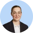 Portrait of a woman with light skin and brown hair pulled back, wearing glasses, a white collared shirt, and a dark blazer, against a light blue background.