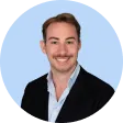 Young man with short brown hair and mustache wearing a black blazer and white shirt, smiling against a pale blue circular background.