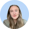 Portrait of a young woman with long brown hair, wearing a gray blazer, smiling against a light blue circular background.