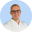 Smiling bald man with glasses wearing a white collared shirt against a light blue background.