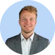 Smiling blond man with short curly hair wearing a gray suit jacket, white shirt, and no tie, against a light blue background in a circular crop.