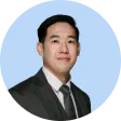 Portrait of an Asian man wearing a dark suit, white shirt, and dark tie, with a light blue circular background.