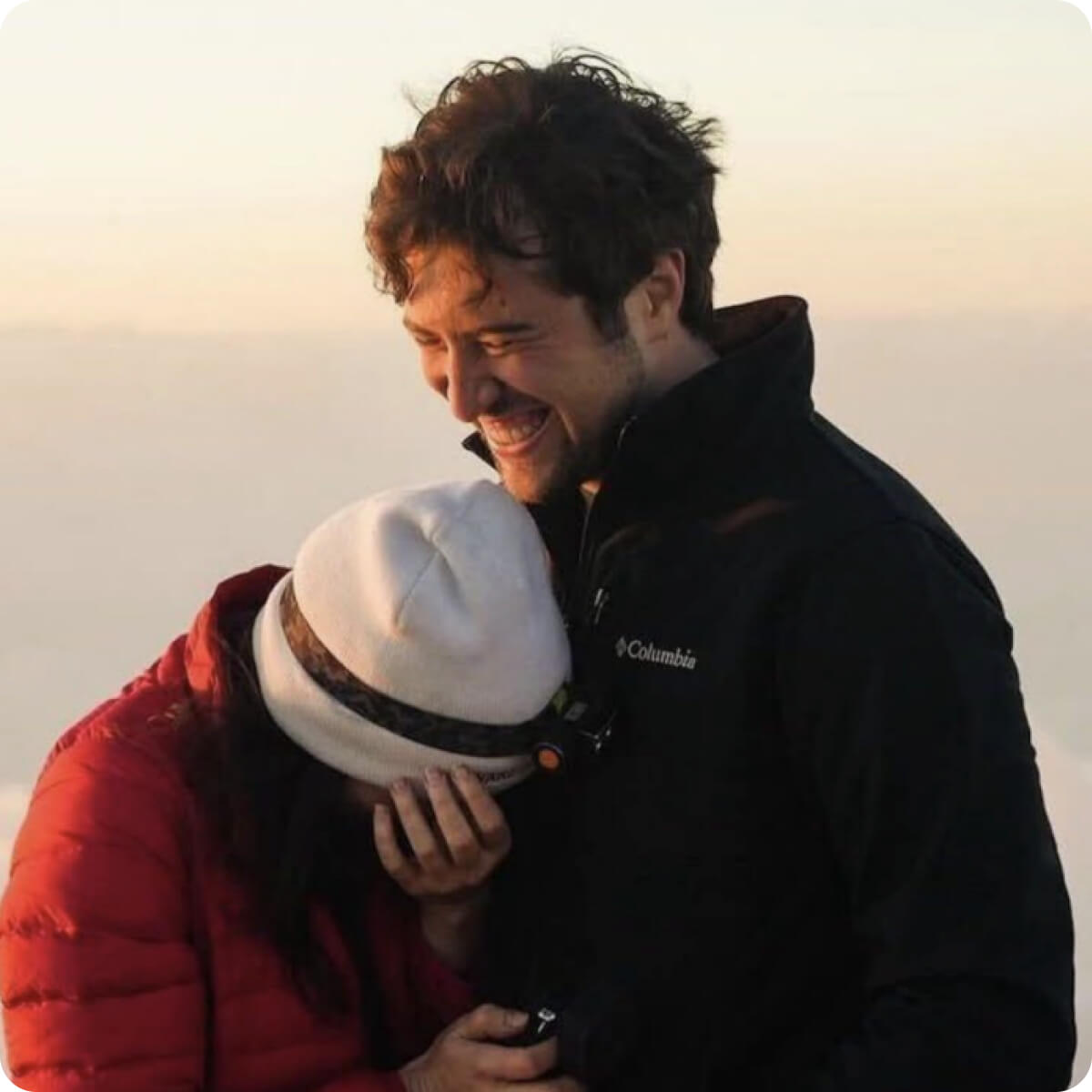 Man in black Columbia jacket smiling warmly as woman in red jacket and white beanie hides her face in his chest.