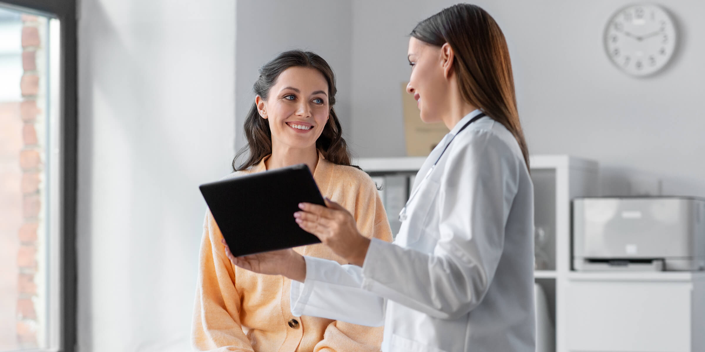 Smiling male doctor with glasses and stethoscope working on a laptop in a bright office.