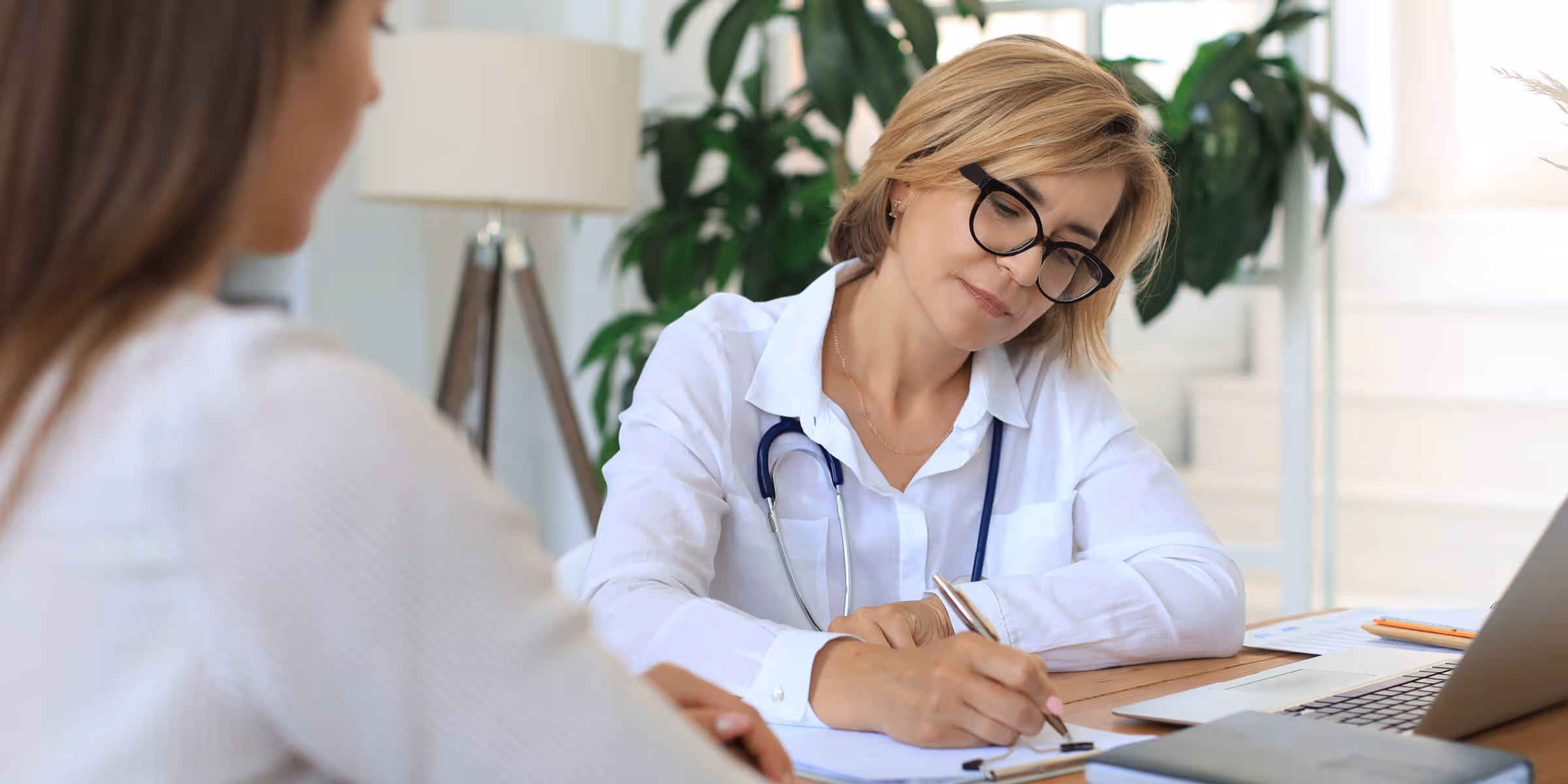Smiling male doctor with glasses and stethoscope working on a laptop in a bright office.