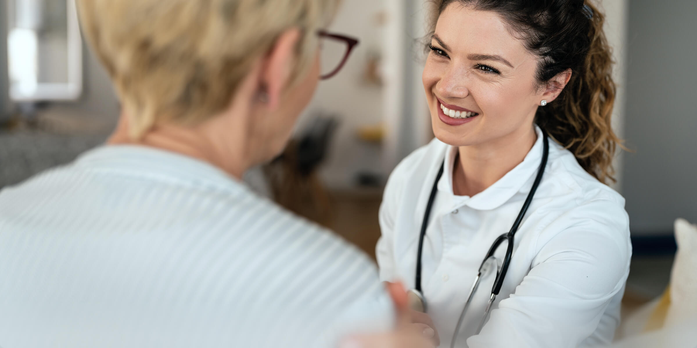 Smiling male doctor with glasses and stethoscope working on a laptop in a bright office.
