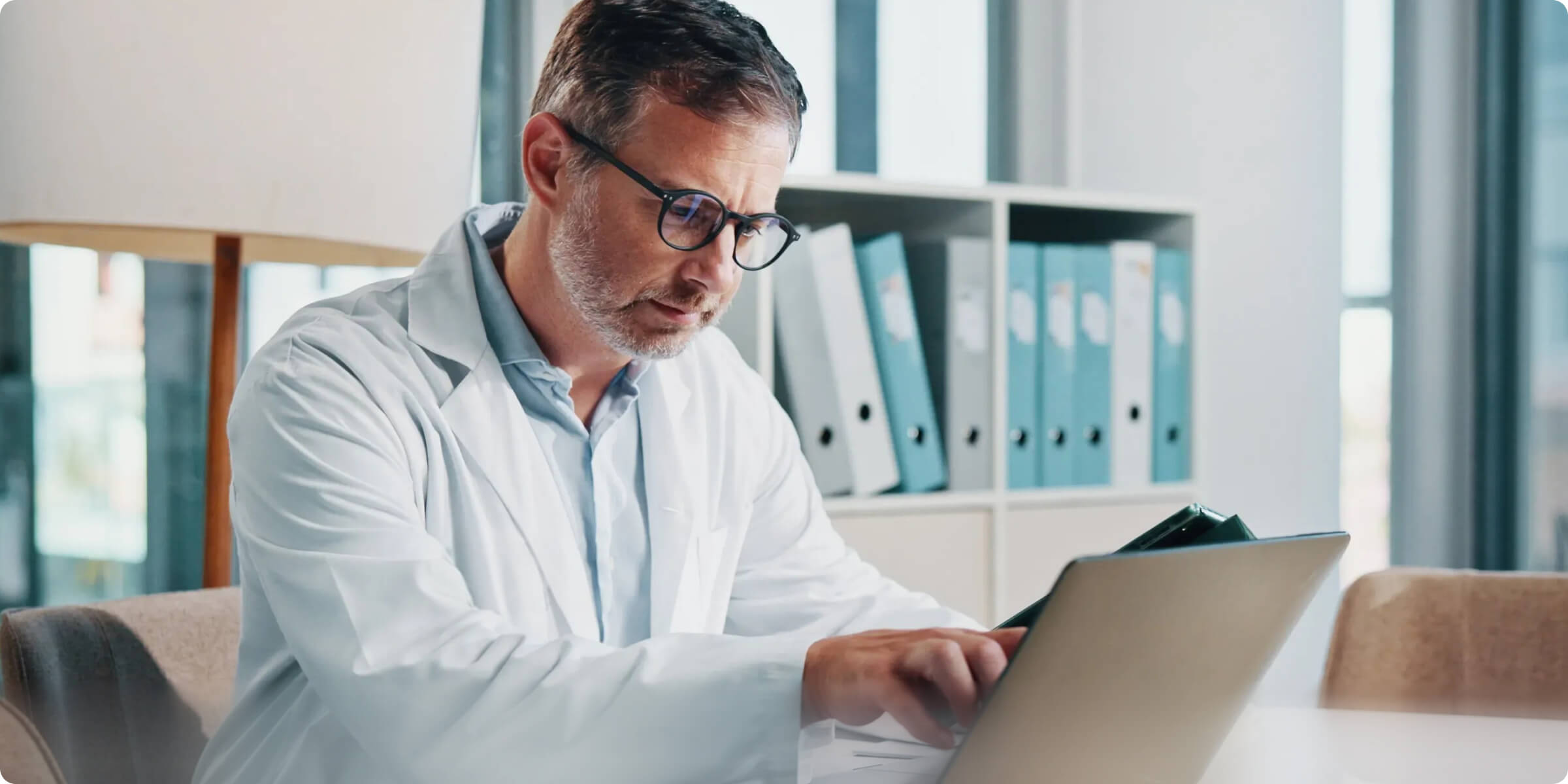 Smiling male doctor with glasses and stethoscope working on a laptop in a bright office.