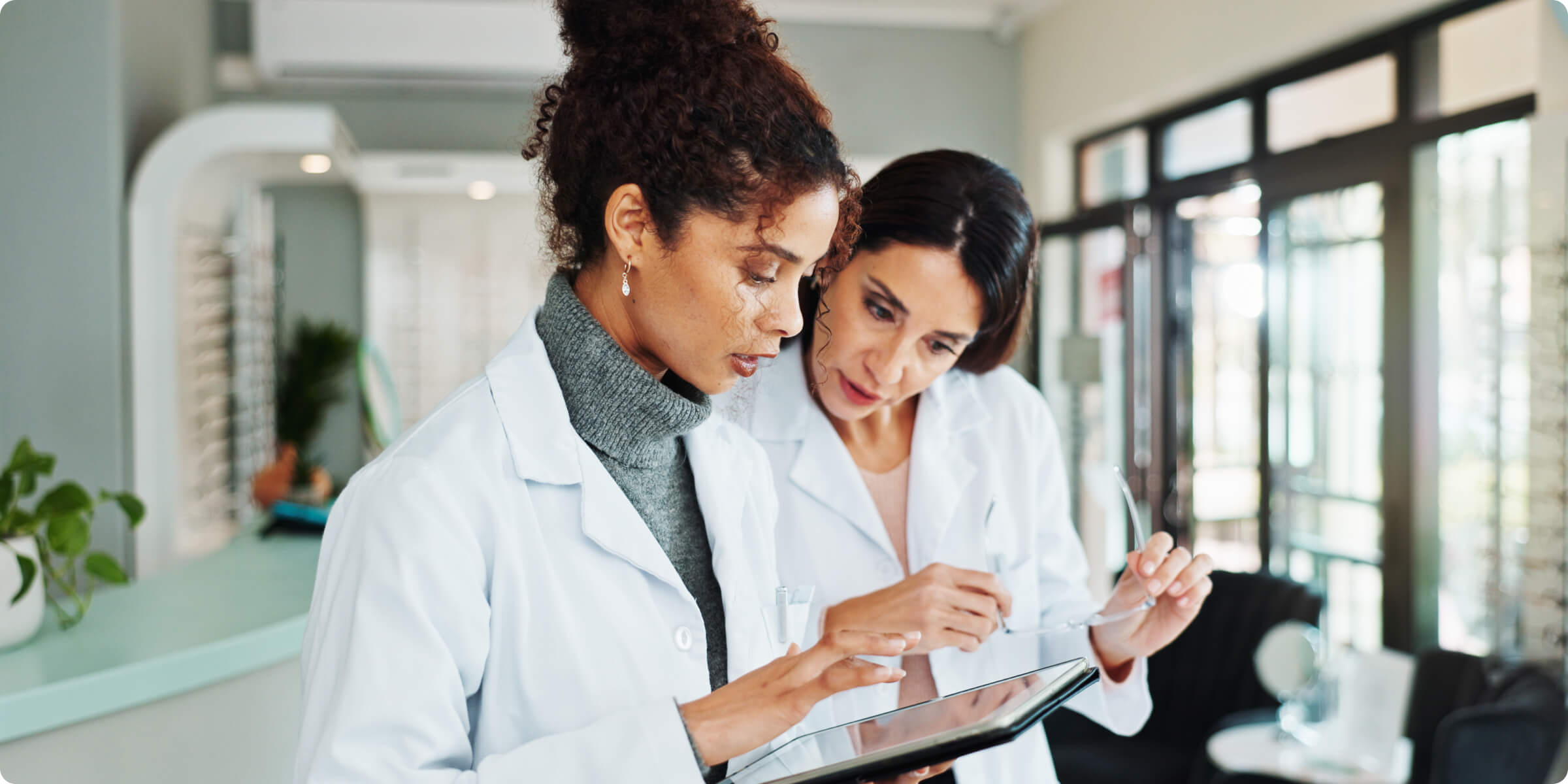 Smiling male doctor with glasses and stethoscope working on a laptop in a bright office.