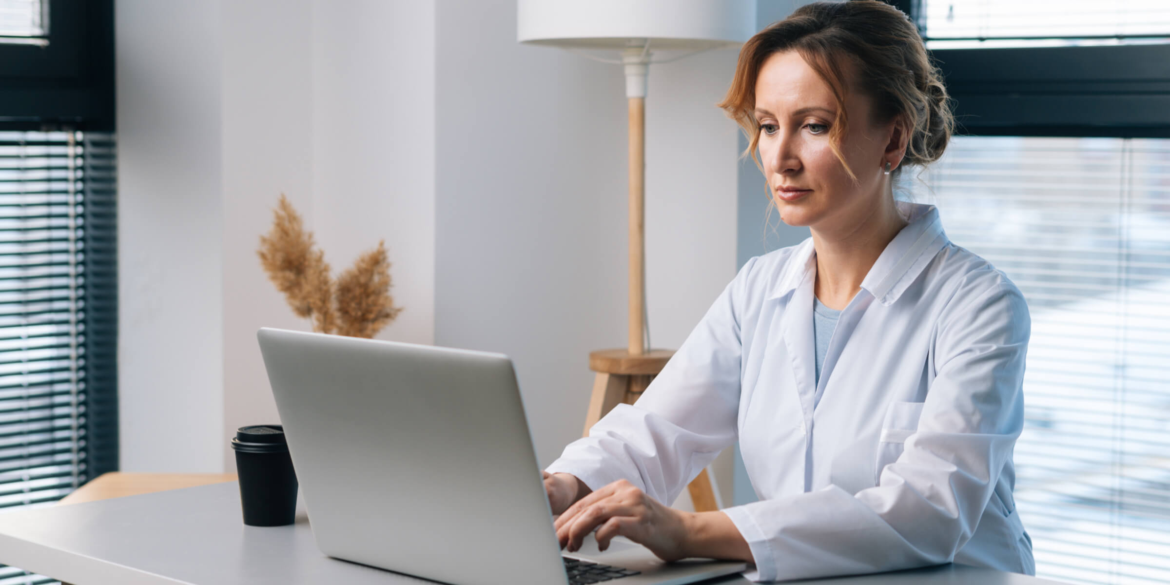 Smiling male doctor with glasses and stethoscope working on a laptop in a bright office.