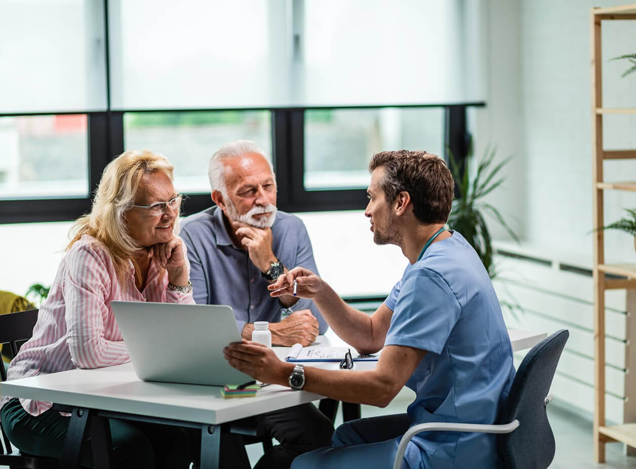 Smiling male healthcare professional in blue scrubs with stethoscope working on a laptop in an office.