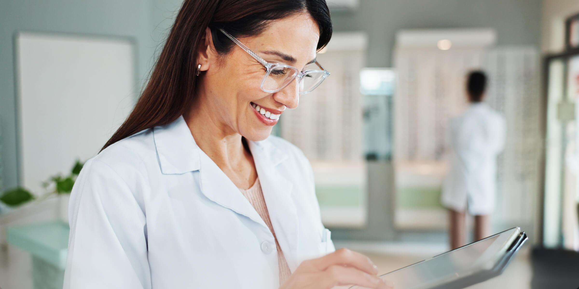 Smiling male doctor with glasses and stethoscope working on a laptop in a bright office.