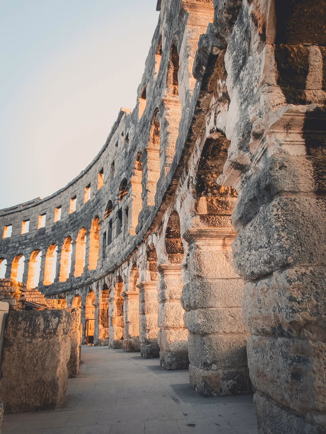 Šikmý pohľad na starovekú rímsku amfiteátr s kameňovými oblúkmi osvetlenými slnečným svetlom.