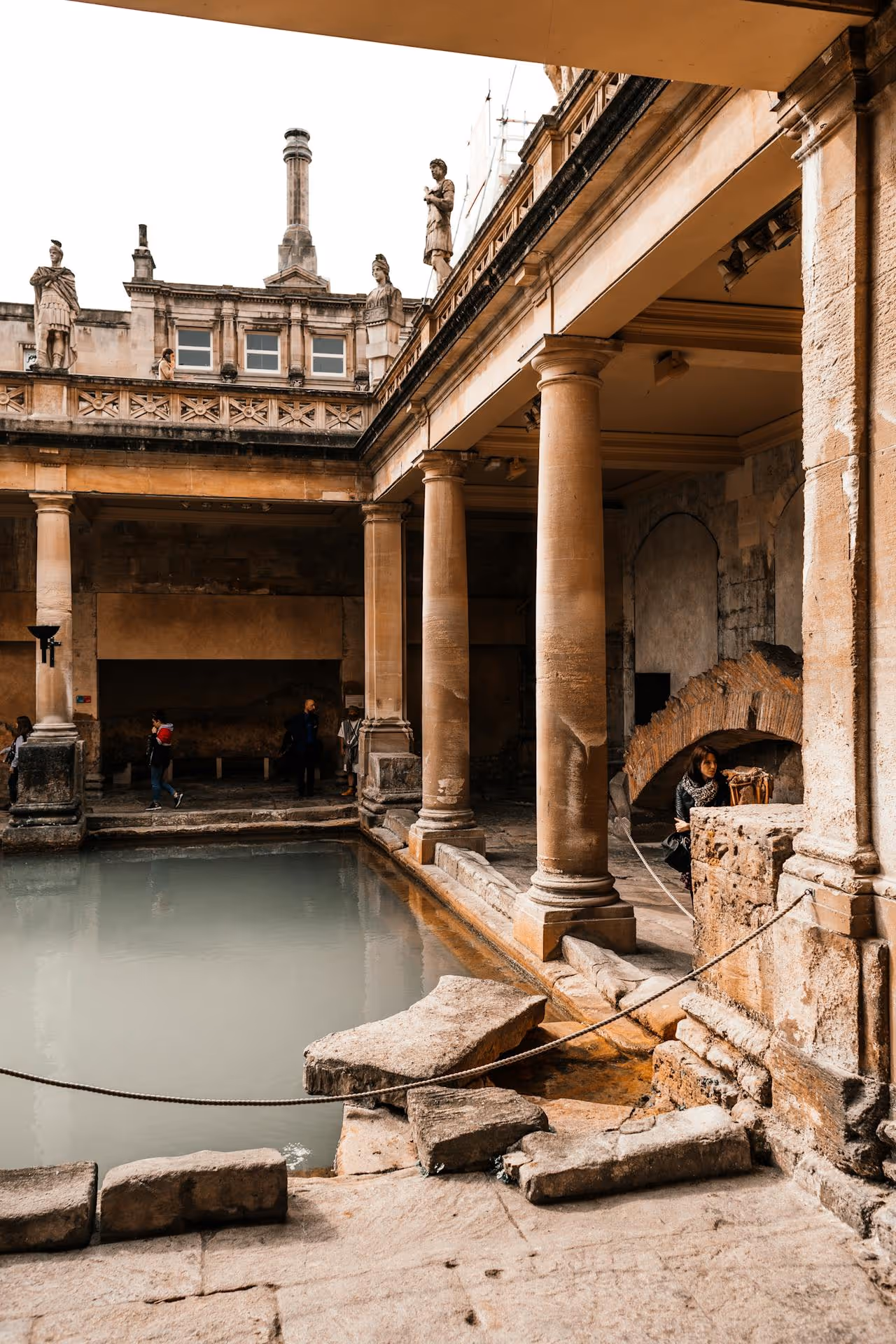 Historické rímske kúpele so stĺpmi, sochami a bazénom zakrištalizovanej vody, kde si ľudia prezerajú a fotografujú pamiatky.