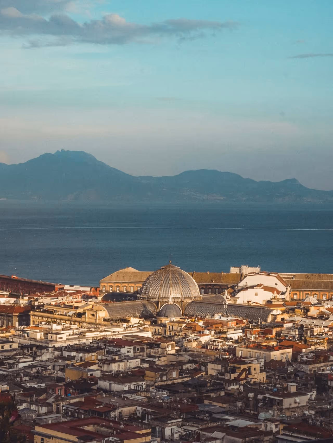 Pohľad na historickú architektúru mesta s kupolovou stavbou pri pobreží a horami v pozadí.