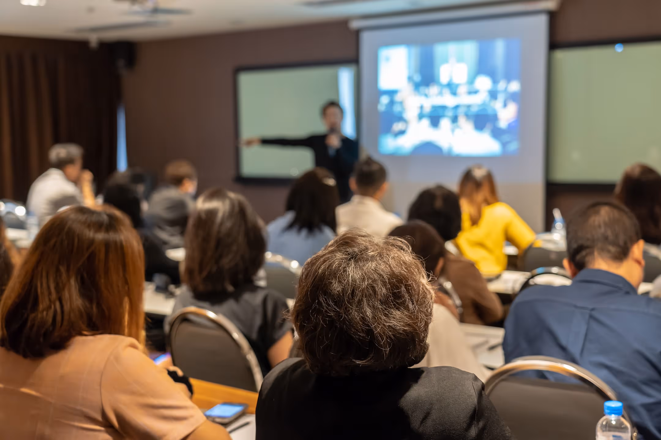 Audience seated in a classroom facing a speaker presenting with a projection screen.