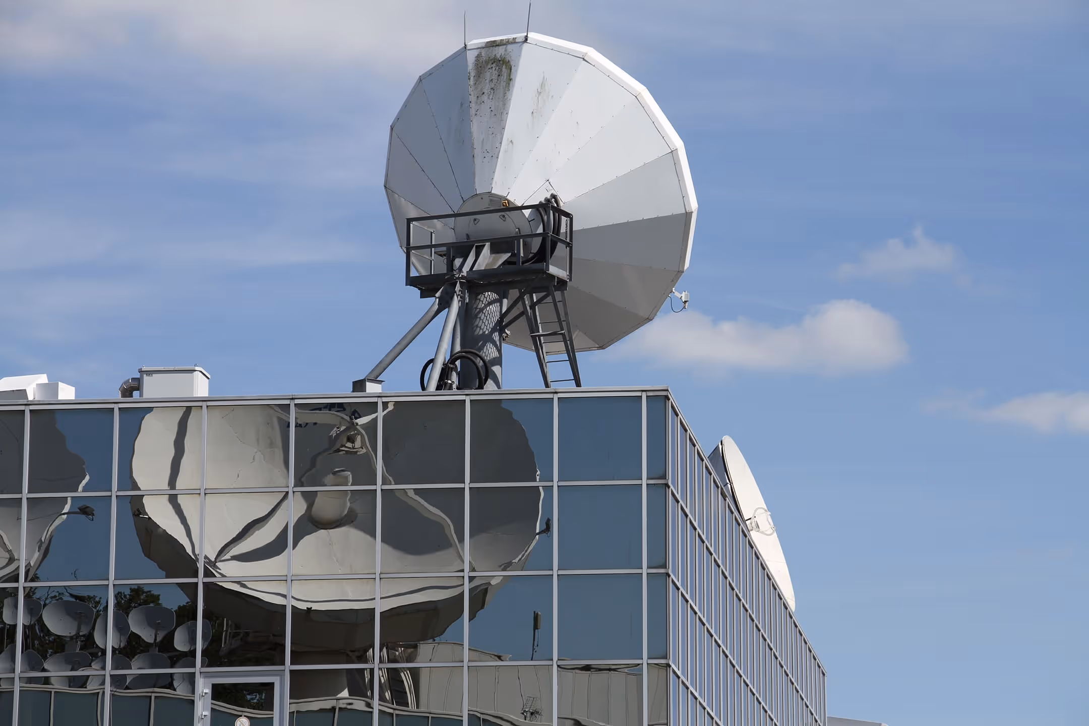 Large white satellite dish mounted on a modern glass building under a blue sky.
