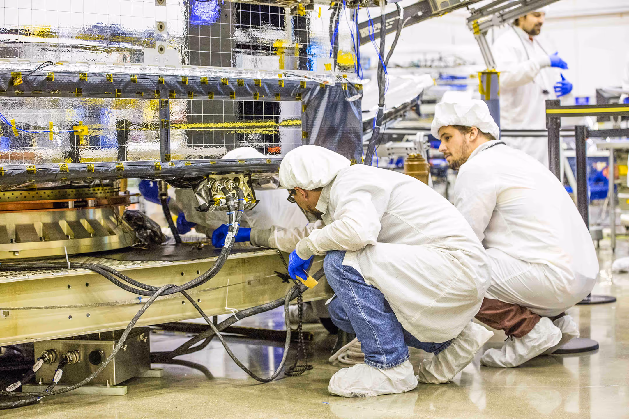 Technicians in cleanroom attire inspecting and working on a large satellite component with wiring and reflective surfaces in a manufacturing facility.