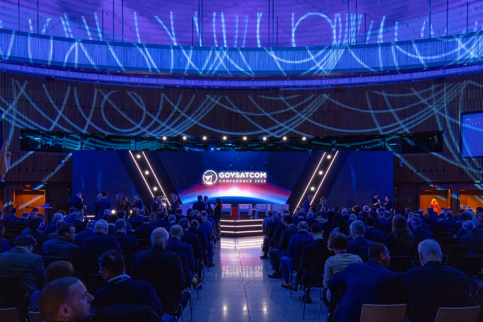 Audience seated facing a stage with a large screen displaying GOVSATCOM Conference 2025 logo and blue swirling light projections on the ceiling.