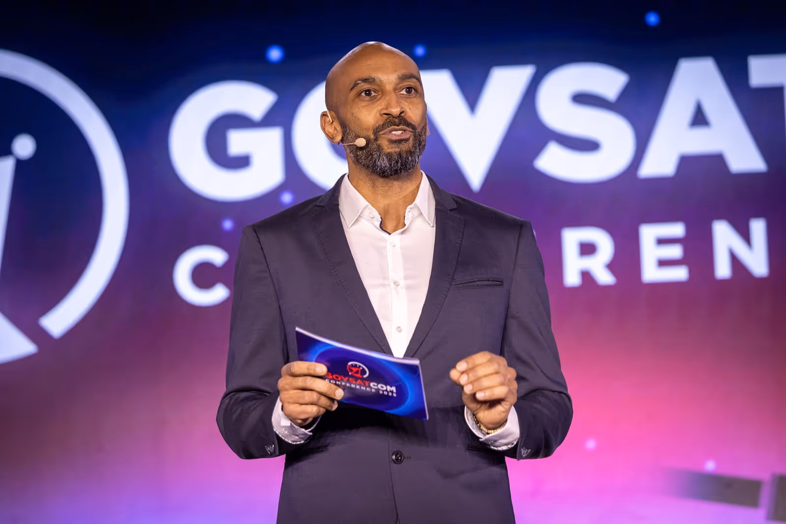 Man in dark suit and white shirt speaking on stage holding a card with GOVSATCOM logo in the background.