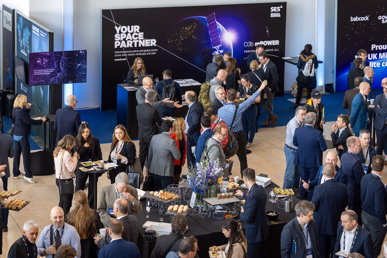 Crowd of business professionals networking and enjoying refreshments at a space industry event with banners and a display booth in the background.