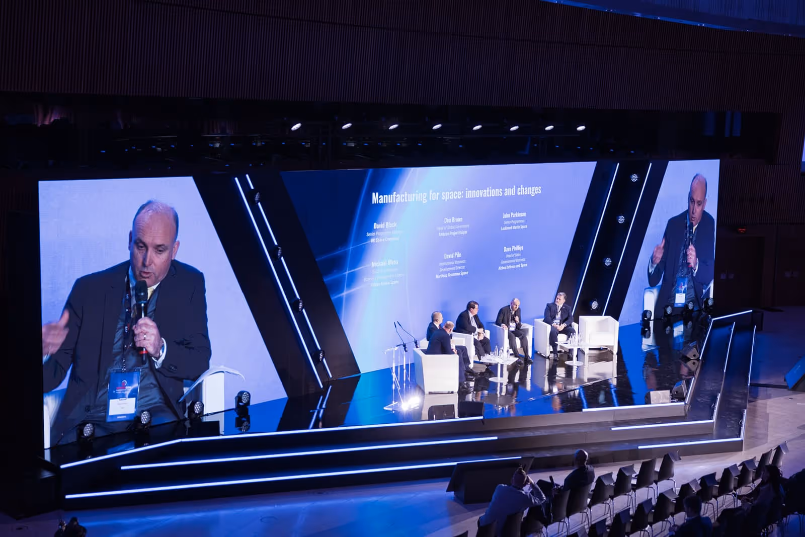 Panel discussion on a stage with large screens showing a man speaking into a microphone and a presentation titled 'Manufacturing for space: innovations and changes'.
