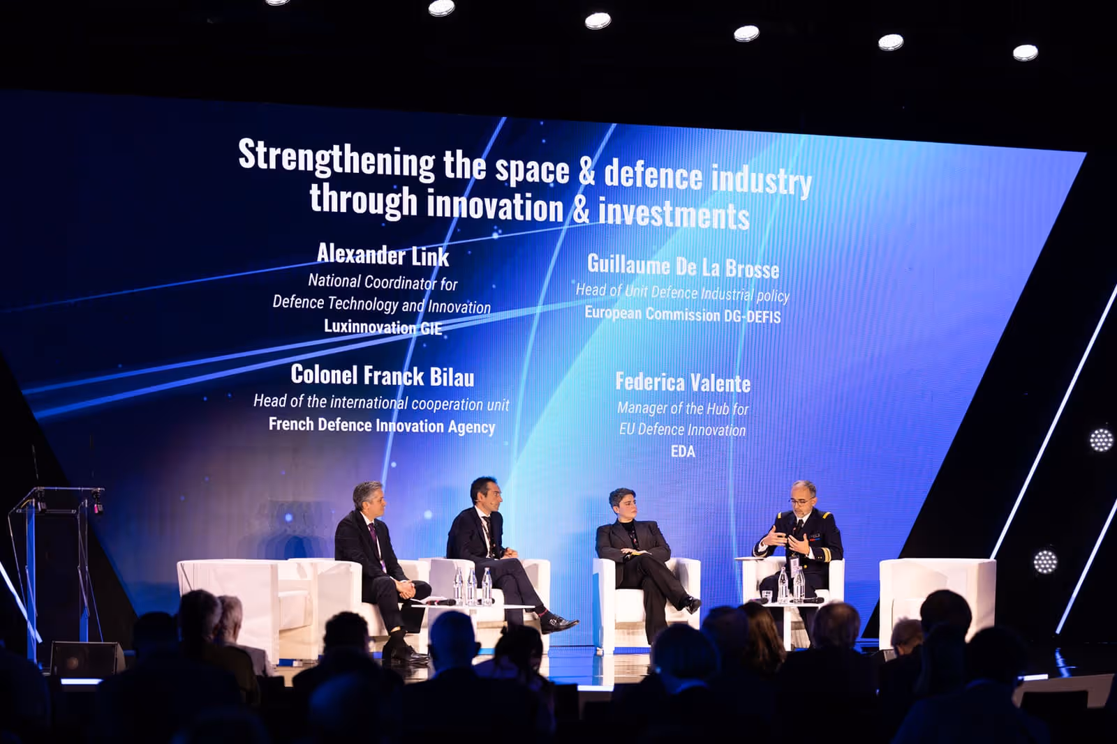 Panel discussion with four speakers seated on white chairs on stage under a large screen displaying the title 'Strengthening the space & defence industry through innovation & investments' and names of panelists.