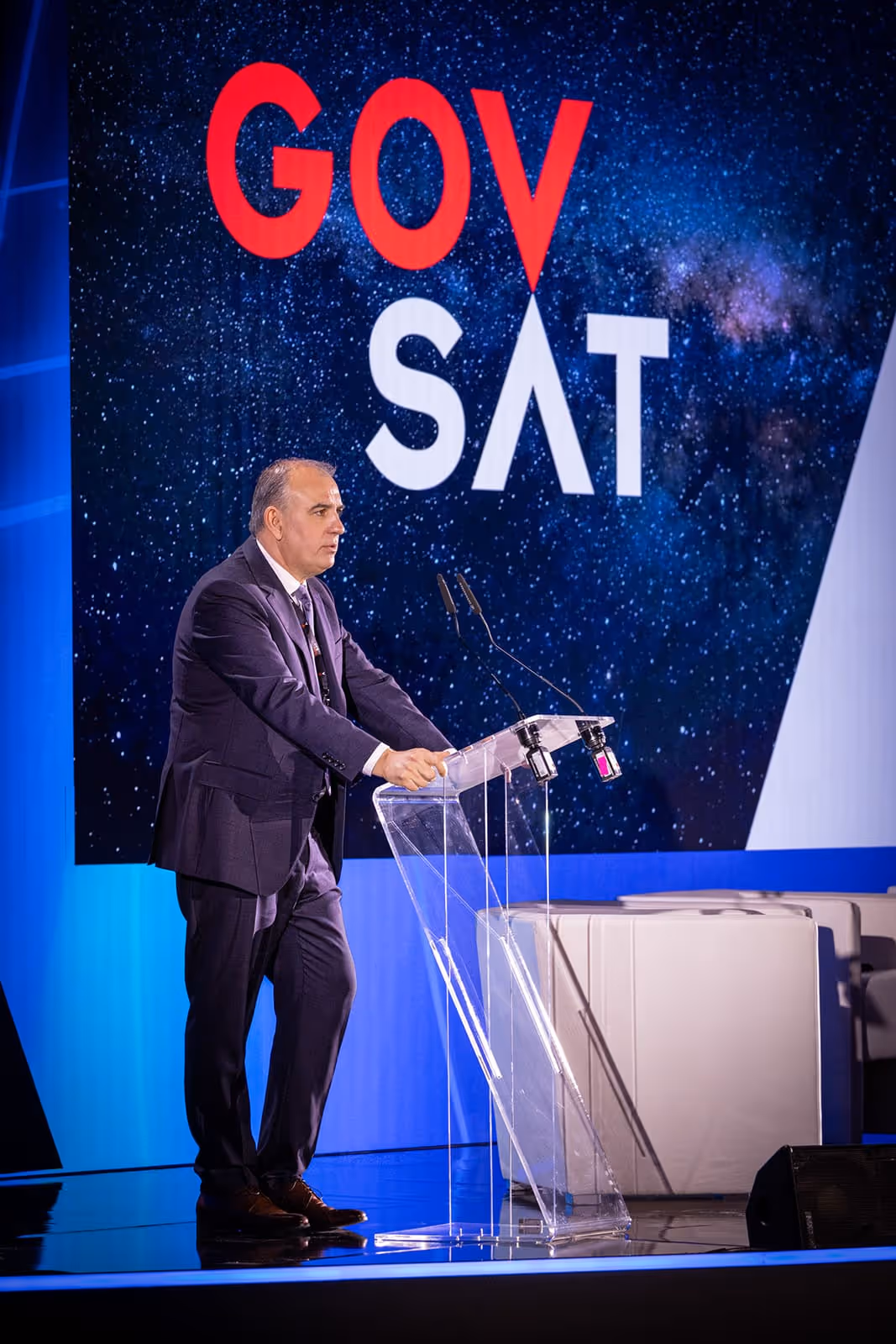 A man in a dark suit stands at a transparent podium speaking, with a large GOVSAT logo and starry sky backdrop behind him.