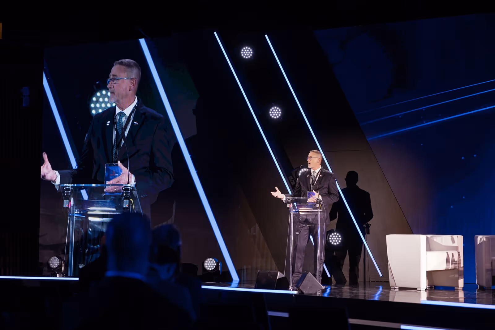 Man in a suit speaking at a clear podium on a stage with diagonal blue light strips and large screen behind him.