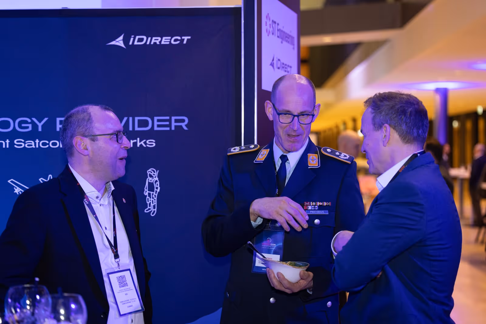 Three men in conversation at an event, including one in a uniform with insignia and two in business attire.