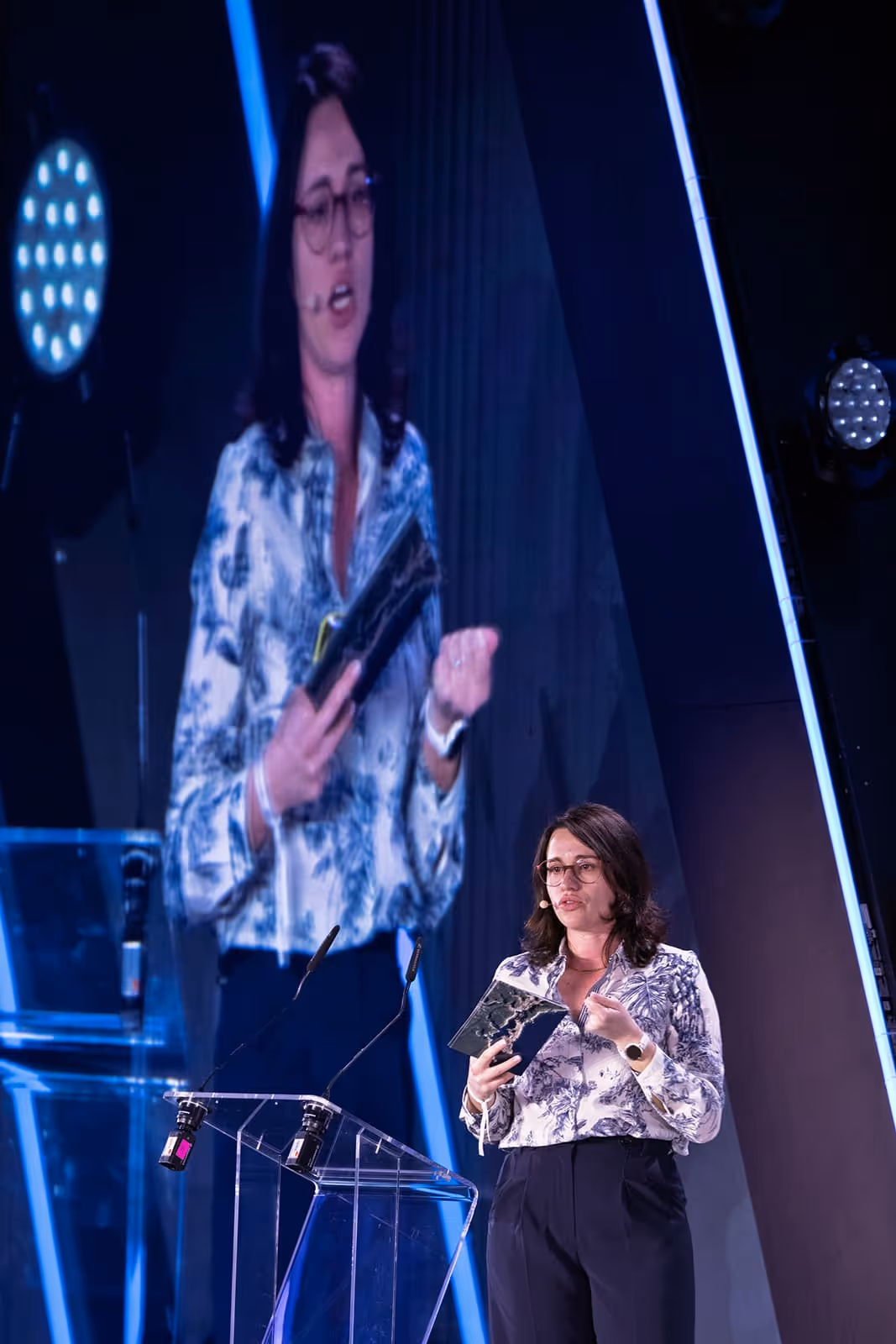 Woman with glasses speaking on stage behind a transparent podium, holding a notebook, with a large projected image of her in the background.