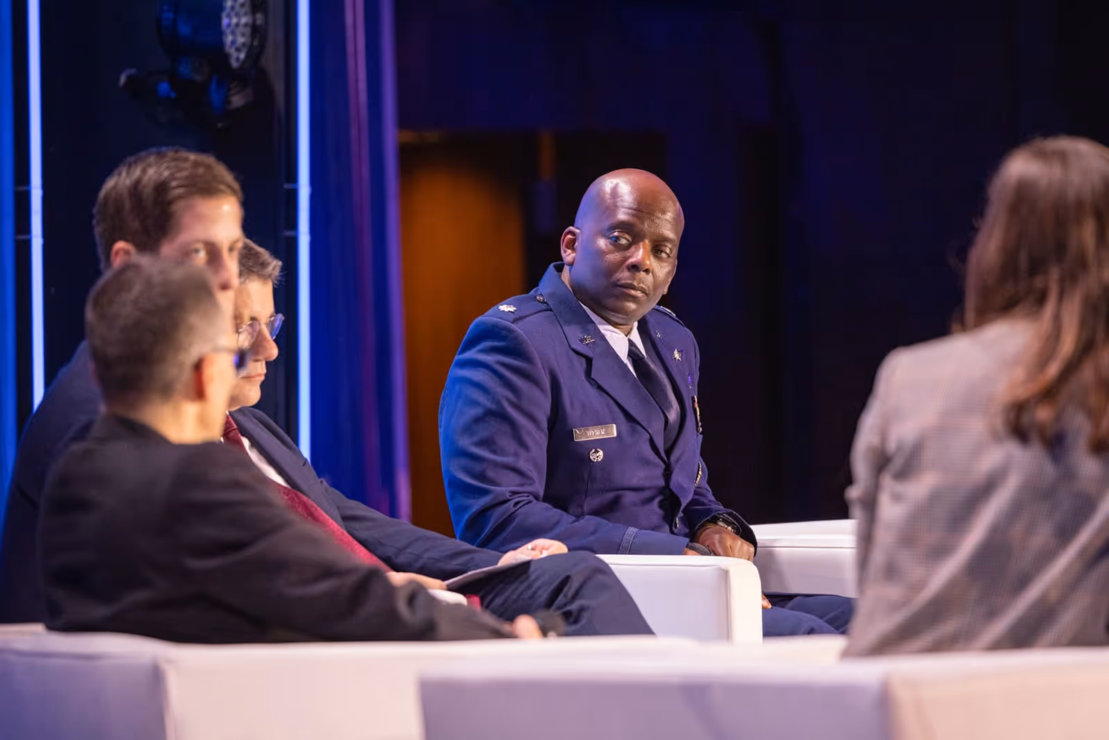 Group of people seated in a panel discussion, including a man in a blue military uniform looking attentively to his right.
