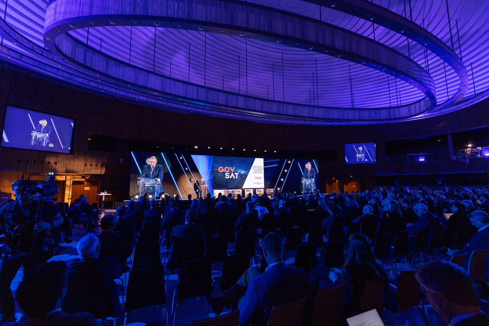 Large audience seated in a dimly lit auditorium watching a speaker at a lectern on stage with GOVSAT branding on screens.