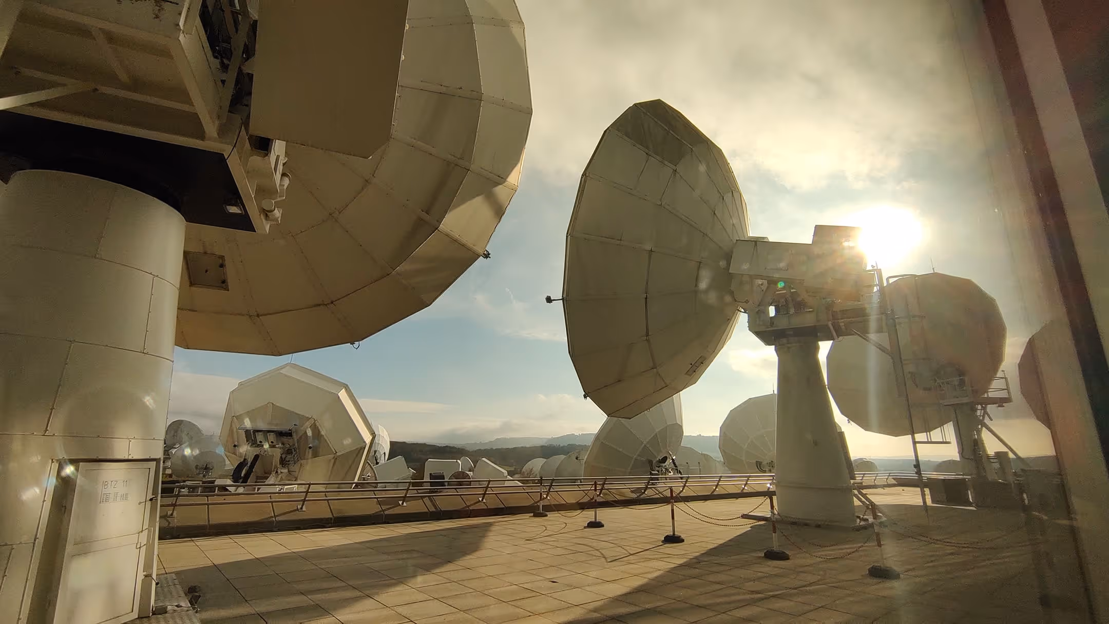 Satellite communication dishes on a rooftop under a bright sky with the sun shining through light clouds.