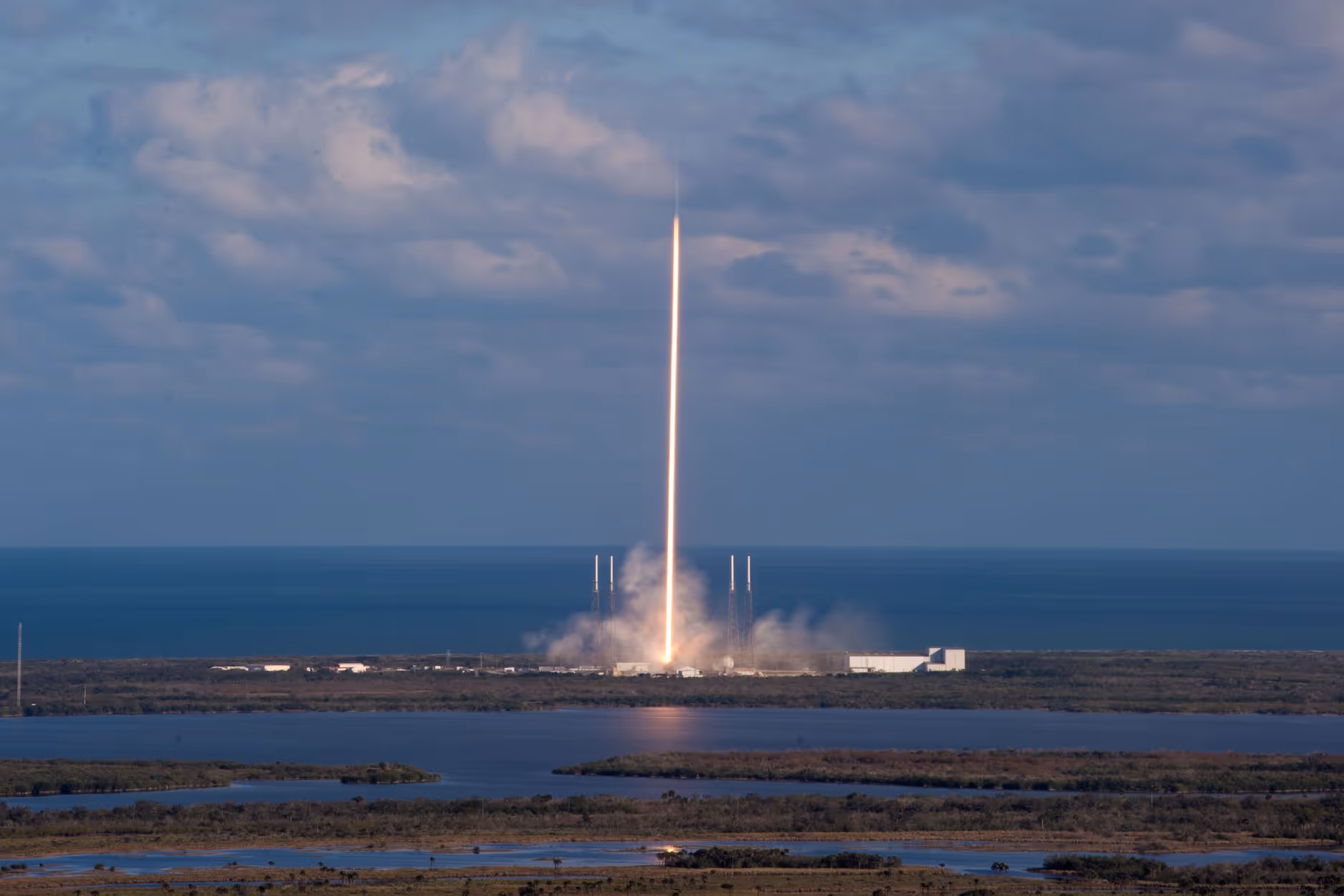 Rocket launching vertically from a coastal launch site with smoke at the base and the ocean in the background.