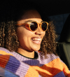 Smiling woman with curly hair wearing orange sunglasses and a colorful striped sweater in sunlight.