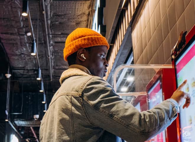 Man wearing an orange beanie and gray jacket uses a touchscreen kiosk indoors.