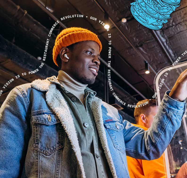 Smiling man wearing an orange beanie and denim jacket with sherpa lining, using a touchscreen device in a modern indoor setting.