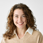 Smiling woman with curly brown hair wearing a beige sweater and white collared shirt.