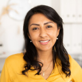 Smiling woman with dark hair wearing a yellow top and pearl earrings in a bright indoor setting.