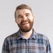 Smiling bearded man wearing a blue and gray plaid shirt against a plain light background.