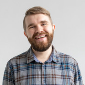 Smiling bearded man wearing a blue and gray plaid shirt against a plain light background.