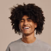 Young man with curly hair laughing with eyes closed against a beige background.
