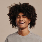 Young man with curly hair laughing with eyes closed against a beige background.