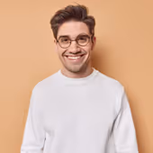 Smiling young man wearing glasses and a white sweater against a beige background.