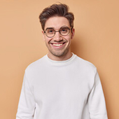 Smiling young man wearing glasses and a white sweater against a beige background.