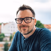 Man with glasses and beard smiling outdoors with blurred houses in background.