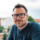 Man with glasses and beard smiling outdoors with blurred houses in background.