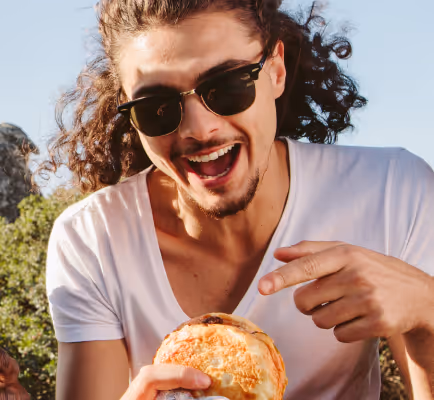 Smiling man with curly hair and sunglasses pointing at a sandwich outdoors.