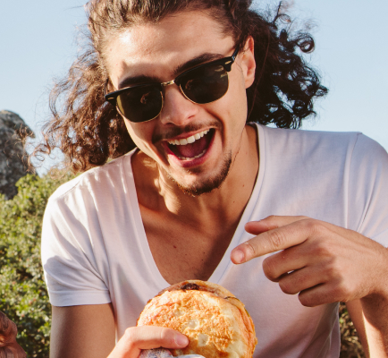 Smiling man with curly hair and sunglasses pointing at a sandwich outdoors.