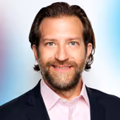 Smiling man with beard and styled brown hair wearing a black blazer and light pink shirt.